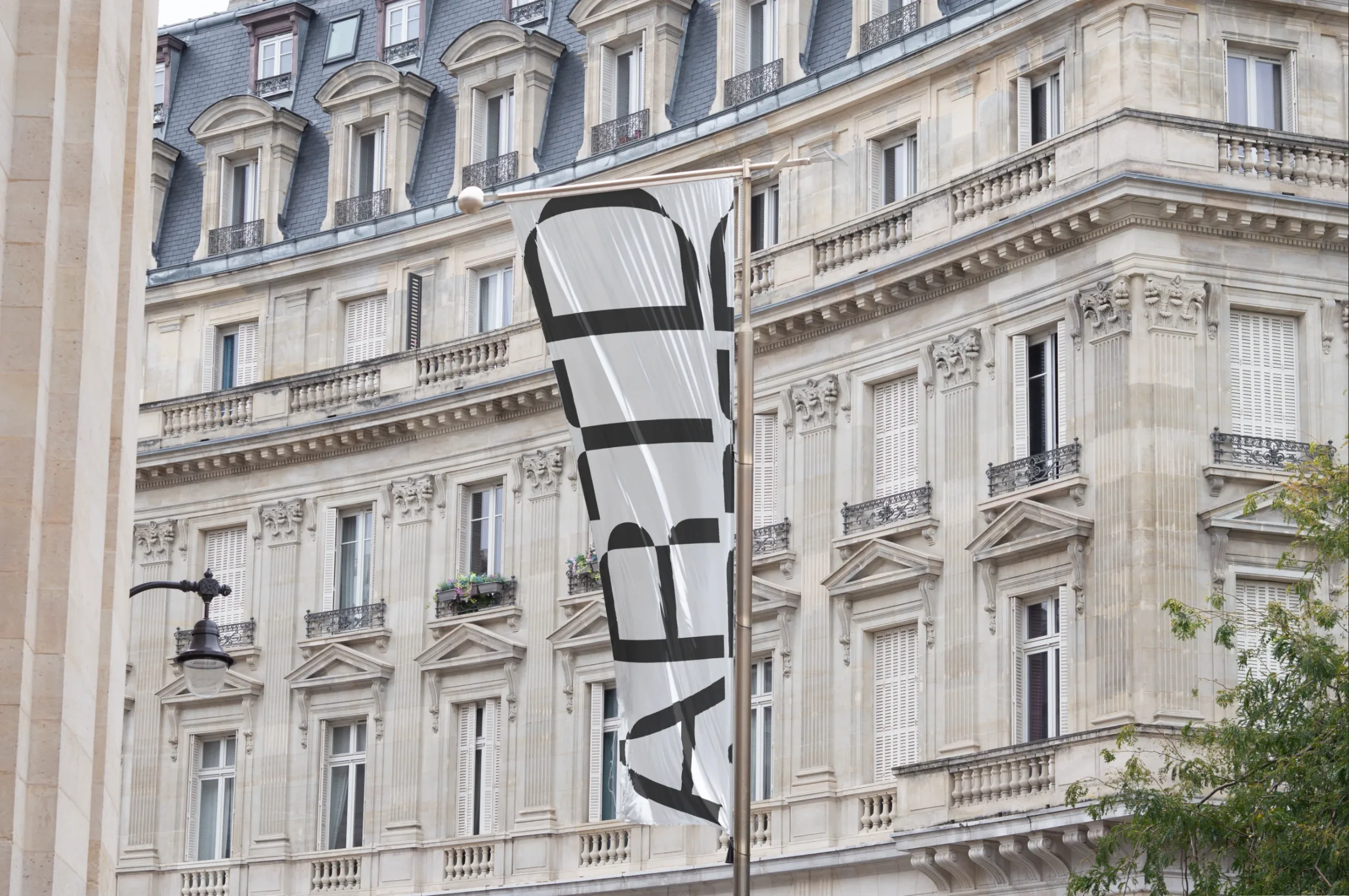 Vertical flag mockup on a street pole featuring a purple banner with bold, abstract black design elements. The background shows an elegant, classical building facade with intricate architectural details.