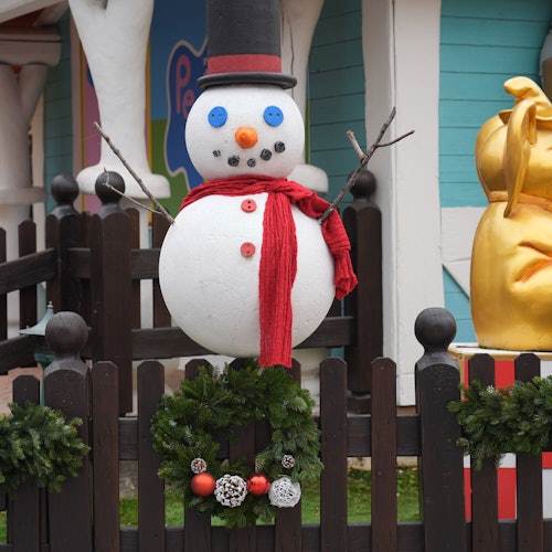 Schneemann mit rotem Schal, Zylinder und Knopfaugen hinter einem schwarzen Zaun, der mit Kränzen geschmückt ist. Rechts befindet sich ein goldenes Objekt.