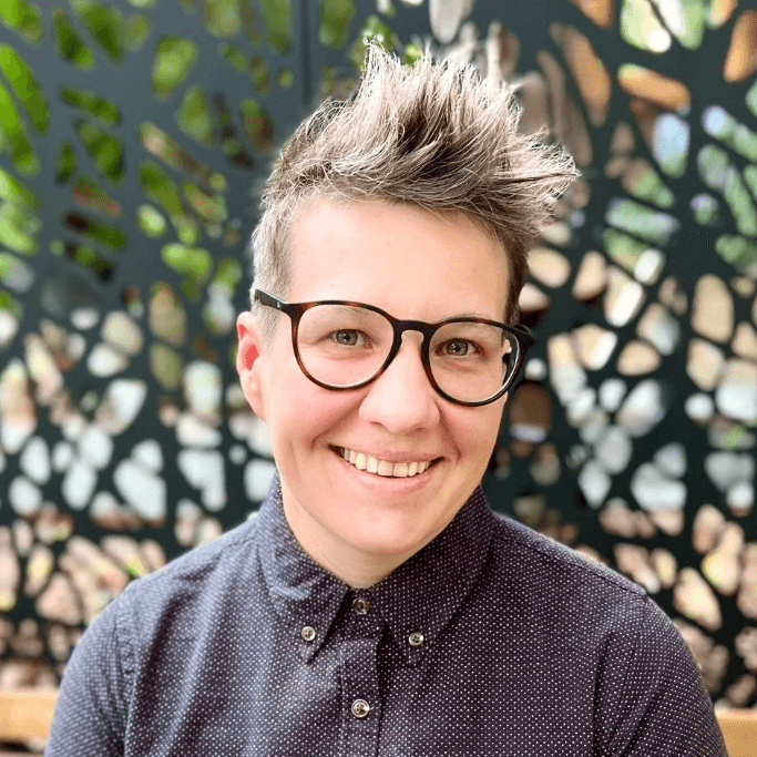 Headshot of Sarah Page, smiling with short, textured hair and round black glasses, wearing a dark button-up shirt, seated in front of a decorative, patterned screen.