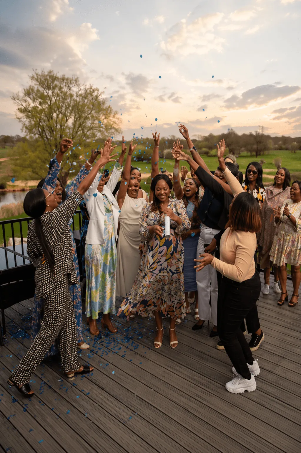 Outdoor gender reveal celebration: joyous group throwing blue confetti,  pregnant woman smiles,  floral dress