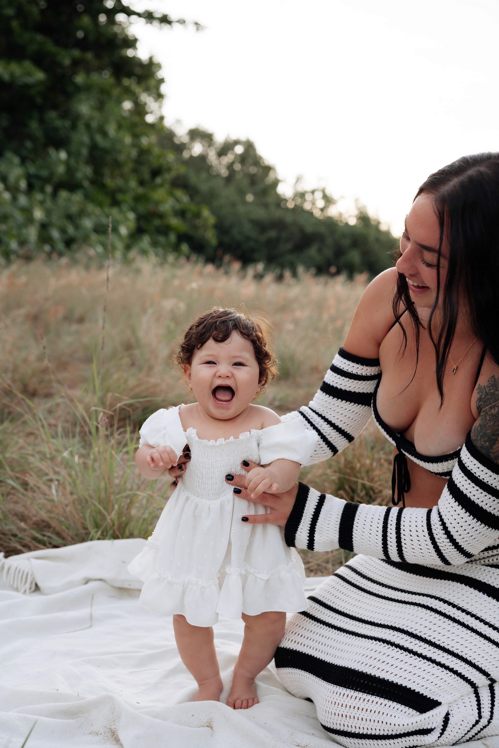 Young child standing beside her mum in tall grass at sunset, captured during a relaxed outdoor motherhood photography session.