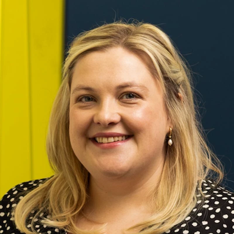A person with blonde hair wears a black polka‑dot top. A bold yellow and dark blue wall provides a colourful backdrop.