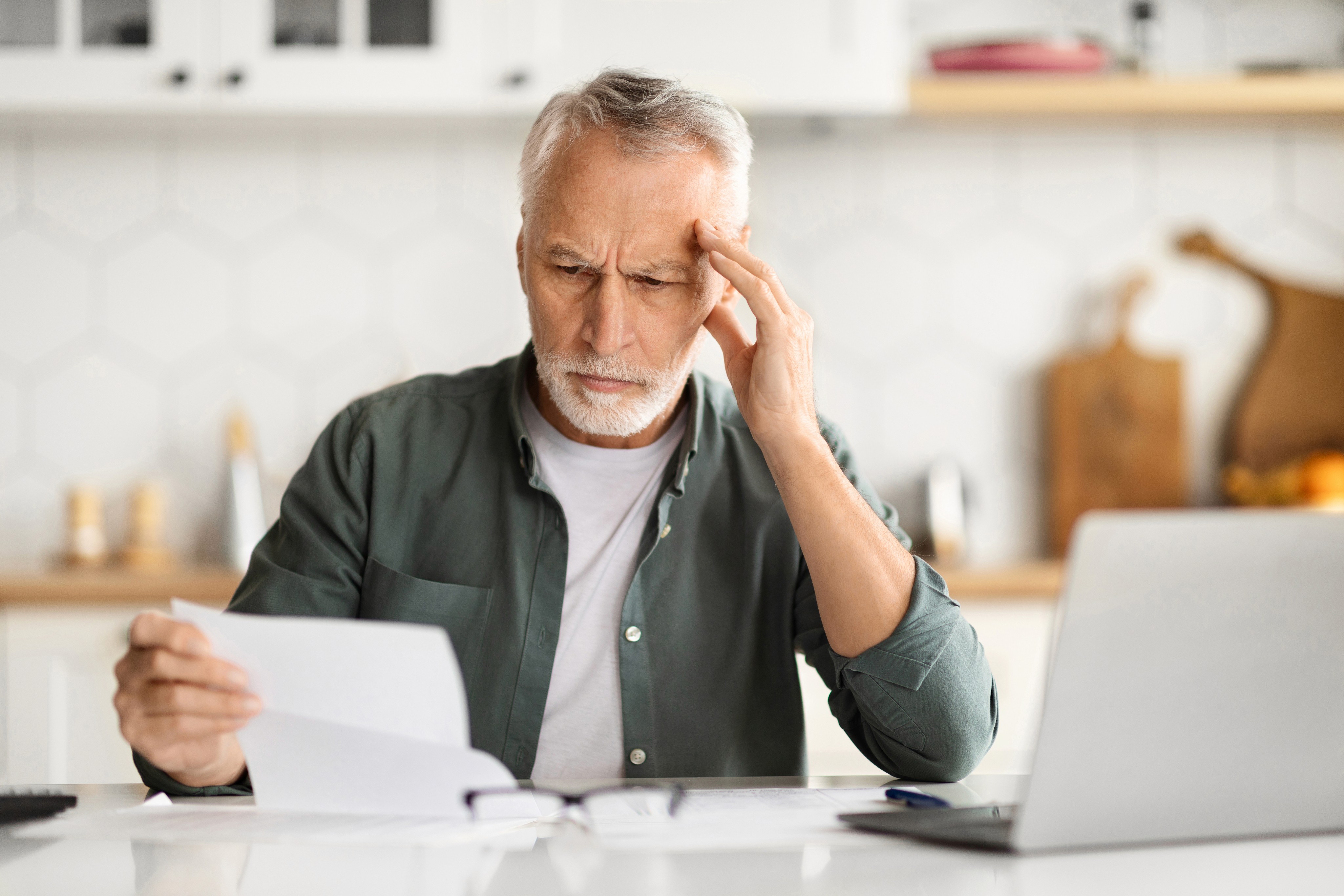 Elderly man with a concerned expression reads documents at a table, resting his hand on his temple. A laptop is open beside him in a bright kitchen.