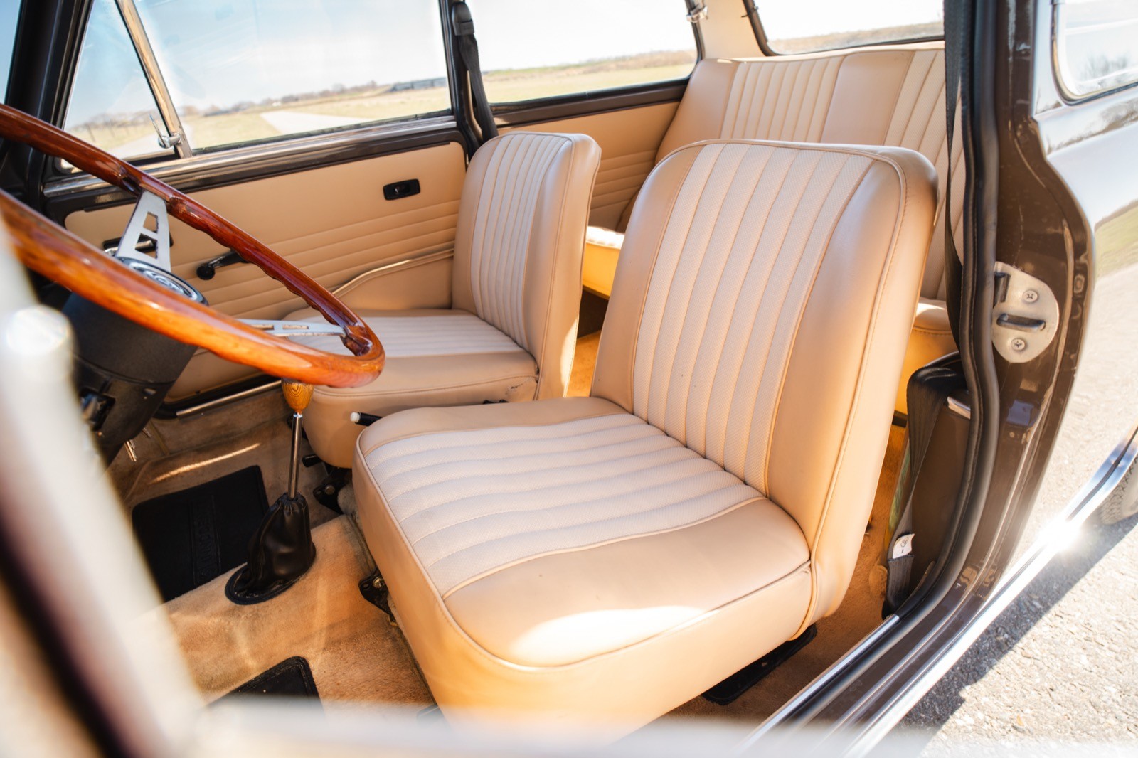 Interior of a car featuring two light-colored leather seats and a wooden dashboard. Bright outdoor light visible.