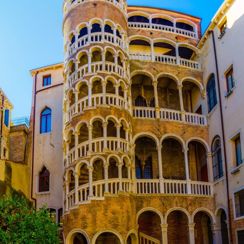 Edificio histórico con una escalera de caracol, varios arcos, balcones y columnas, sobre un cielo azul despejado.