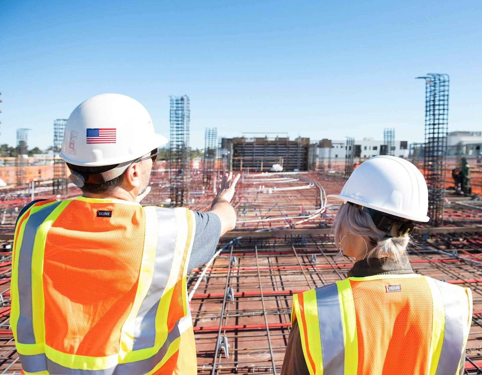 Large commercial construction site with exposed rebar and foundation work, viewed from perspective of two construction professionals in safety gear discussing project progress