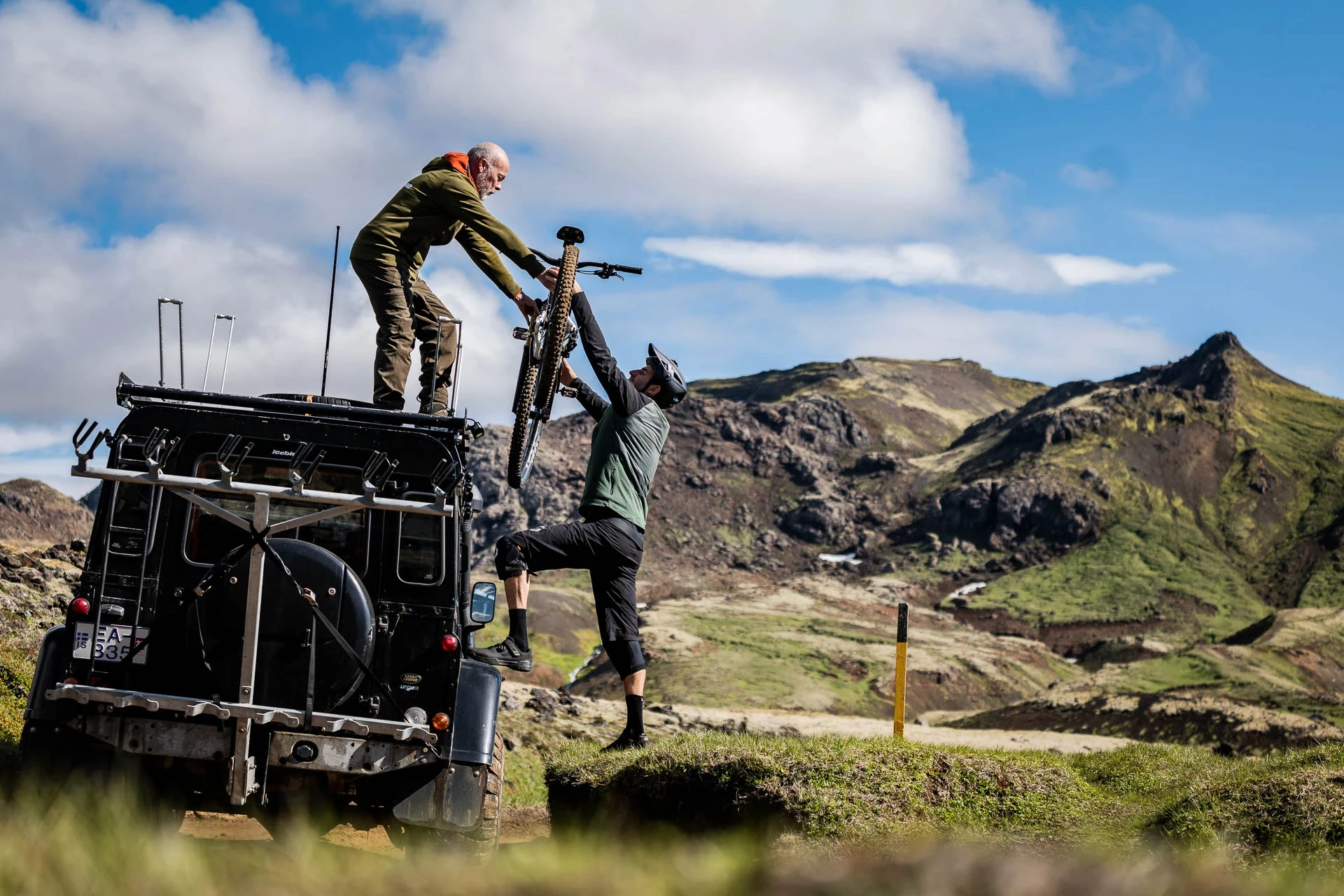 Two people loading a mountain bike onto a 4x4 roof rack with mountains behind.
