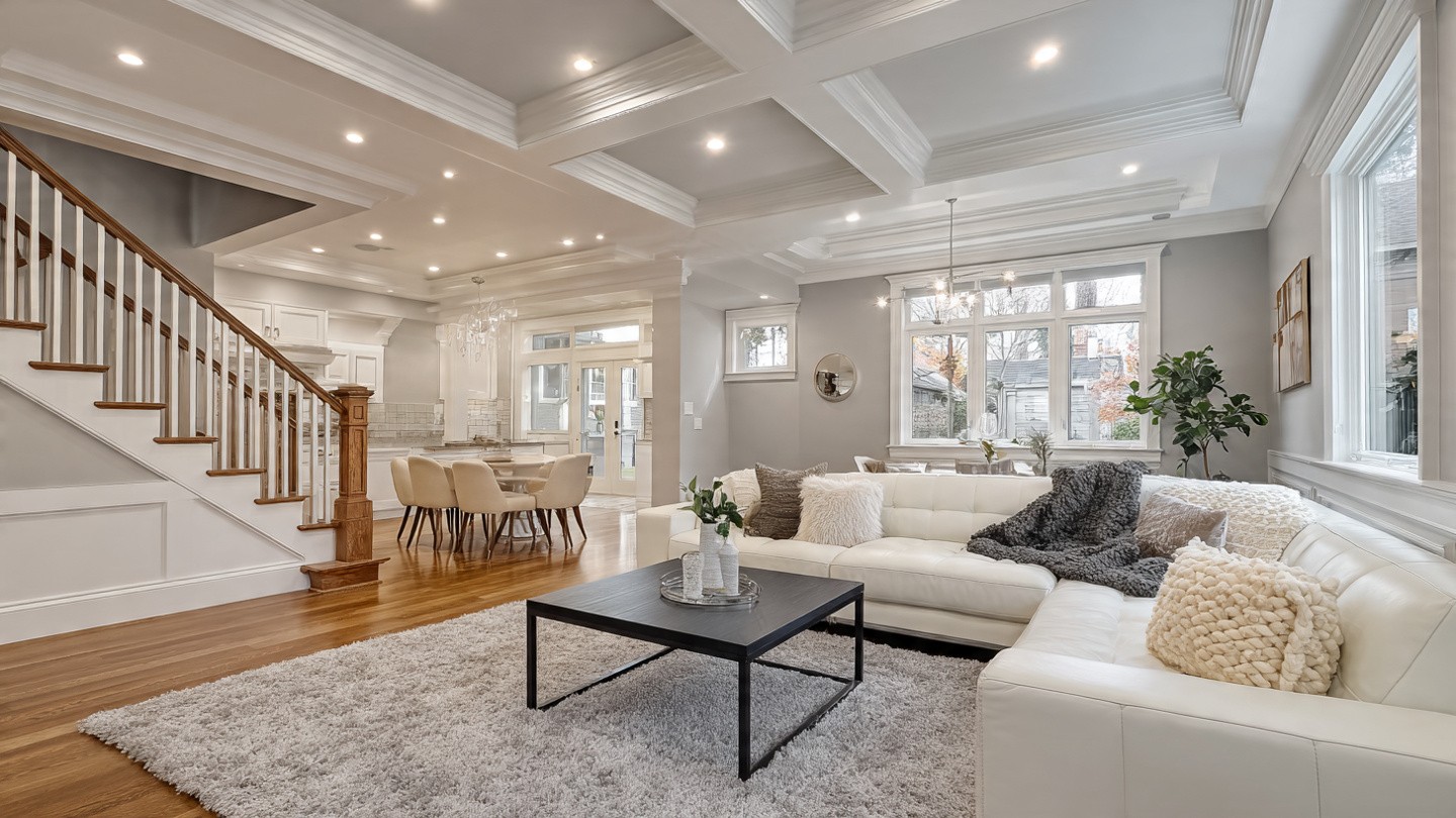 Spacious modern living room with a white sectional sofa, fluffy cushions, and a dark coffee table. Wooden staircase and open kitchen are visible. Cozy and inviting.