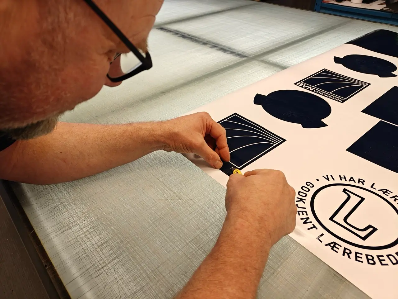 “En person kutter ut klistremerker med ‘Godkjent lærebedrift’-logoen fra en folierull.”