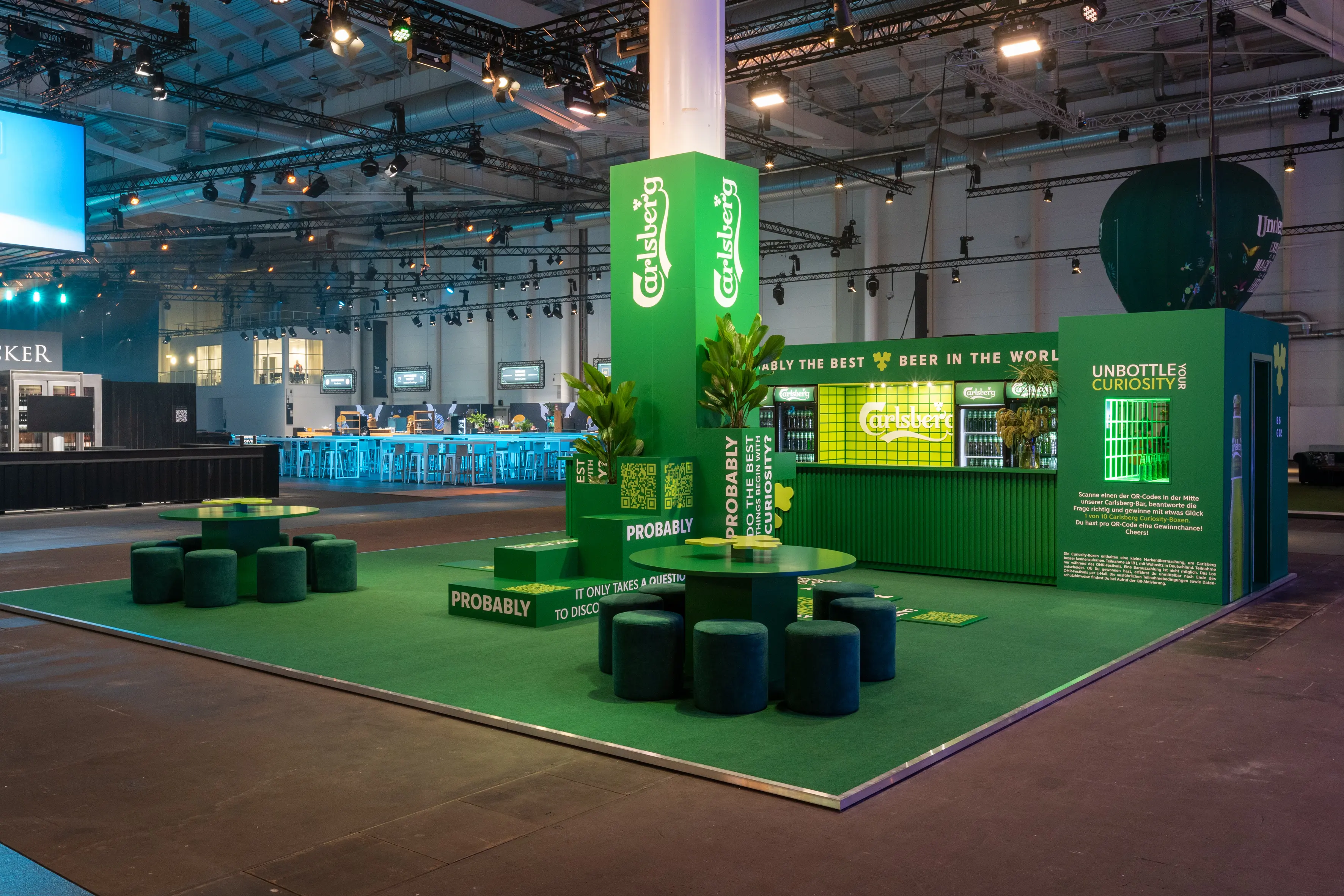 Green Carlsberg trade fair island with logo tower, bar and velvet stools around bistro tables on artificial turf.