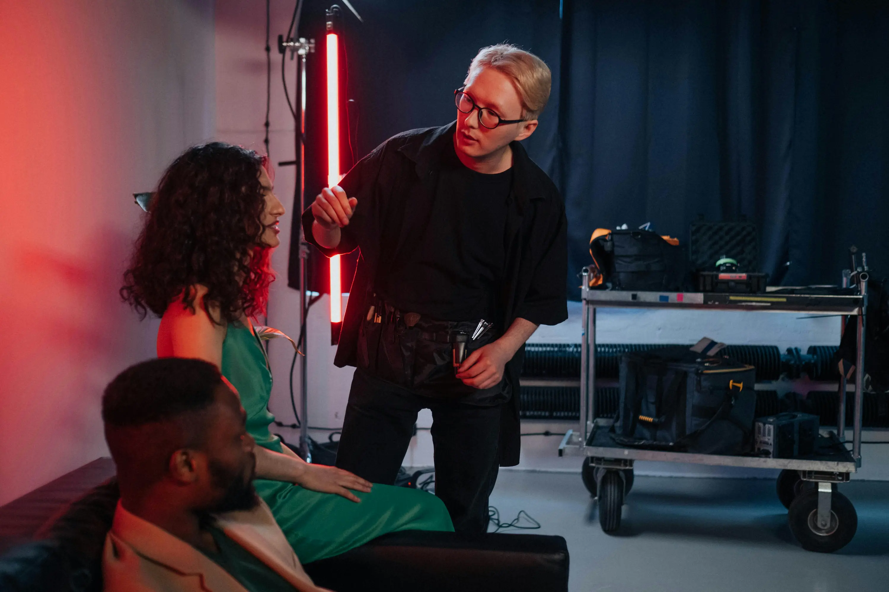 Film crew filming an actor on a couch in a red-lit studio with cameras, a clapperboard, and production lights.