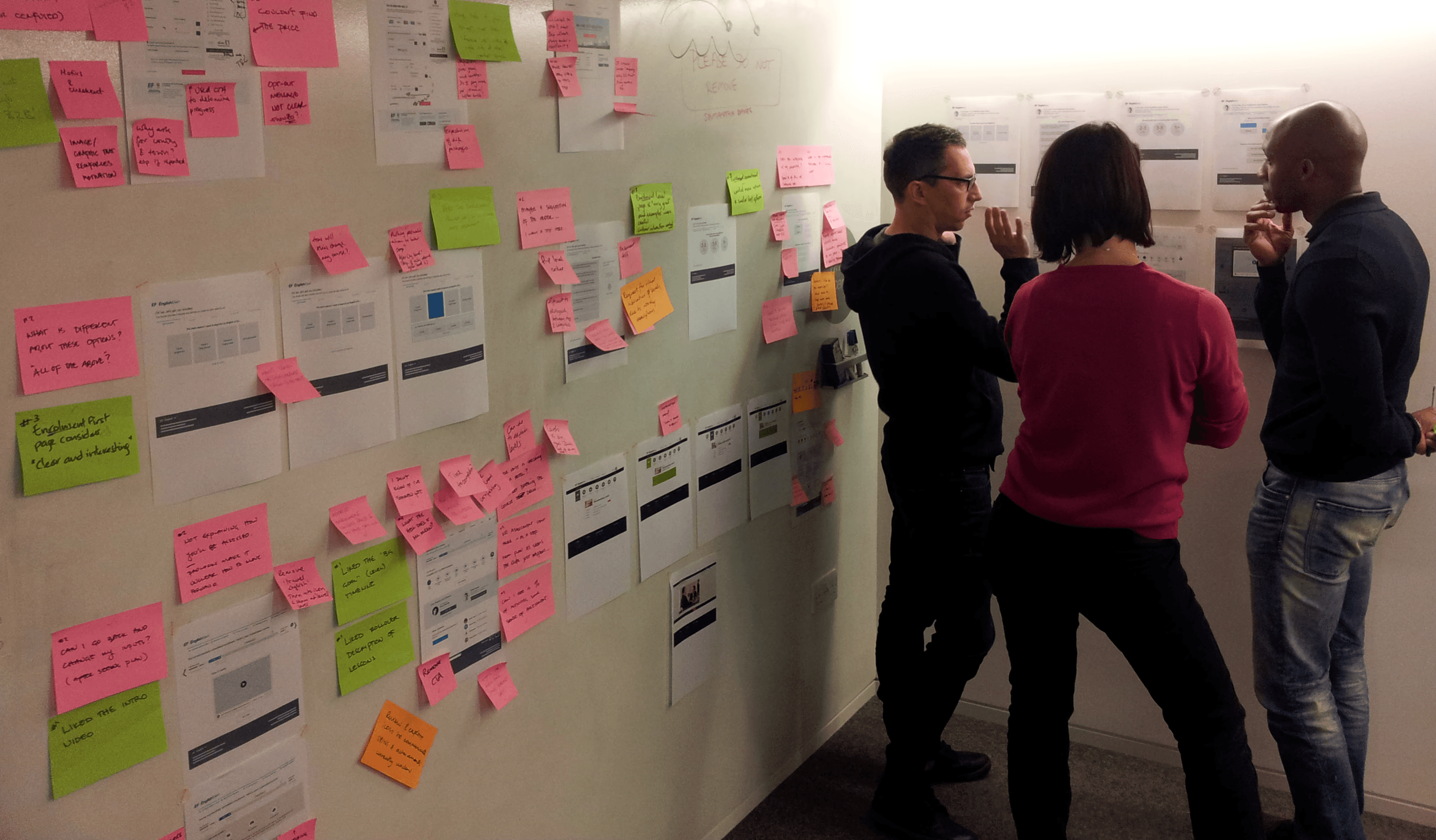 Three people standing at a wall discussing and looking at print-outs of wireframes. The wireframes are annotated with post-its.