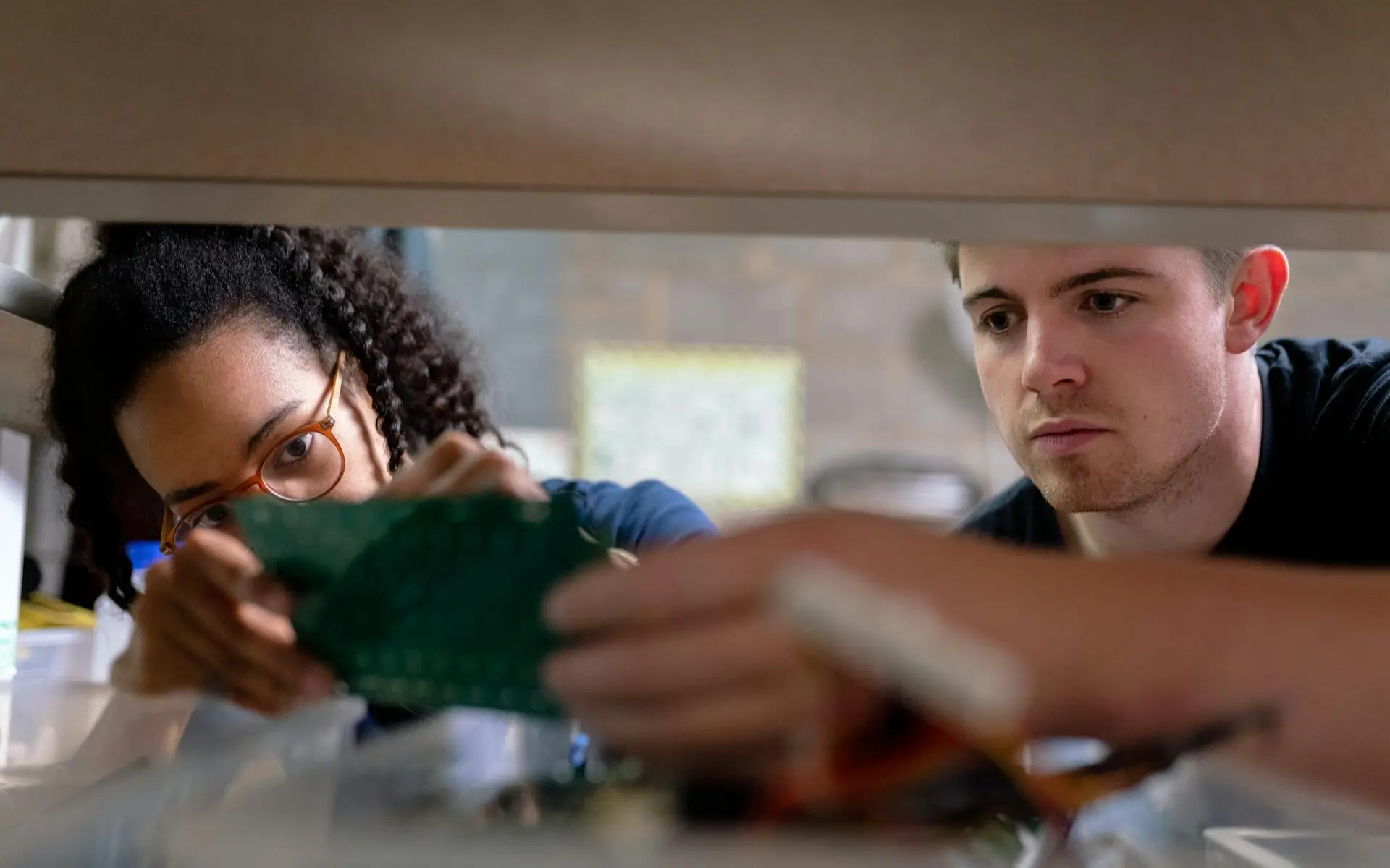 Two people working together on an electronic component, focused on a circuit board with tools visible in the foreground.