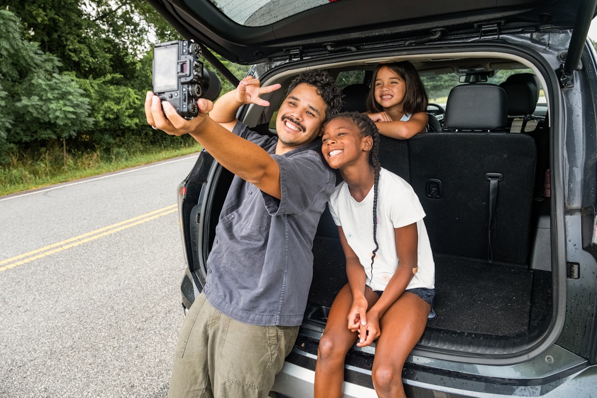 Father and two daughters taking a selfie at an open car tailgate before a road trip
