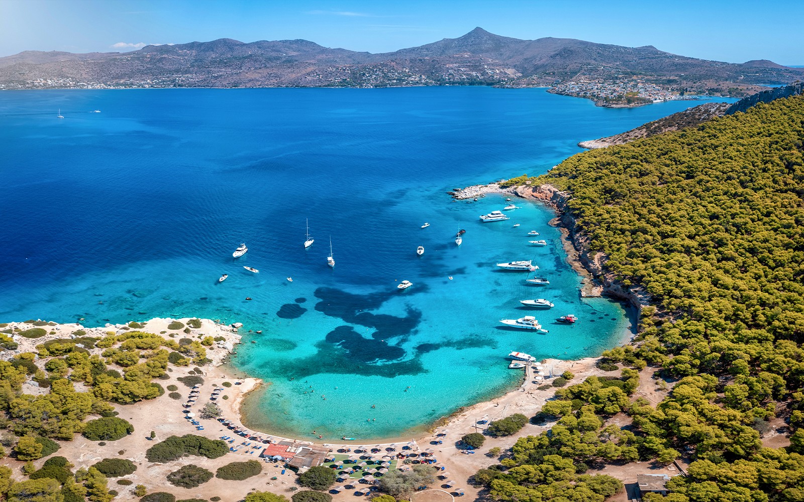 Barche ancorate in una baia turchese vicino ad Atene, circondata da una rigogliosa vegetazione e montagne lontane.
