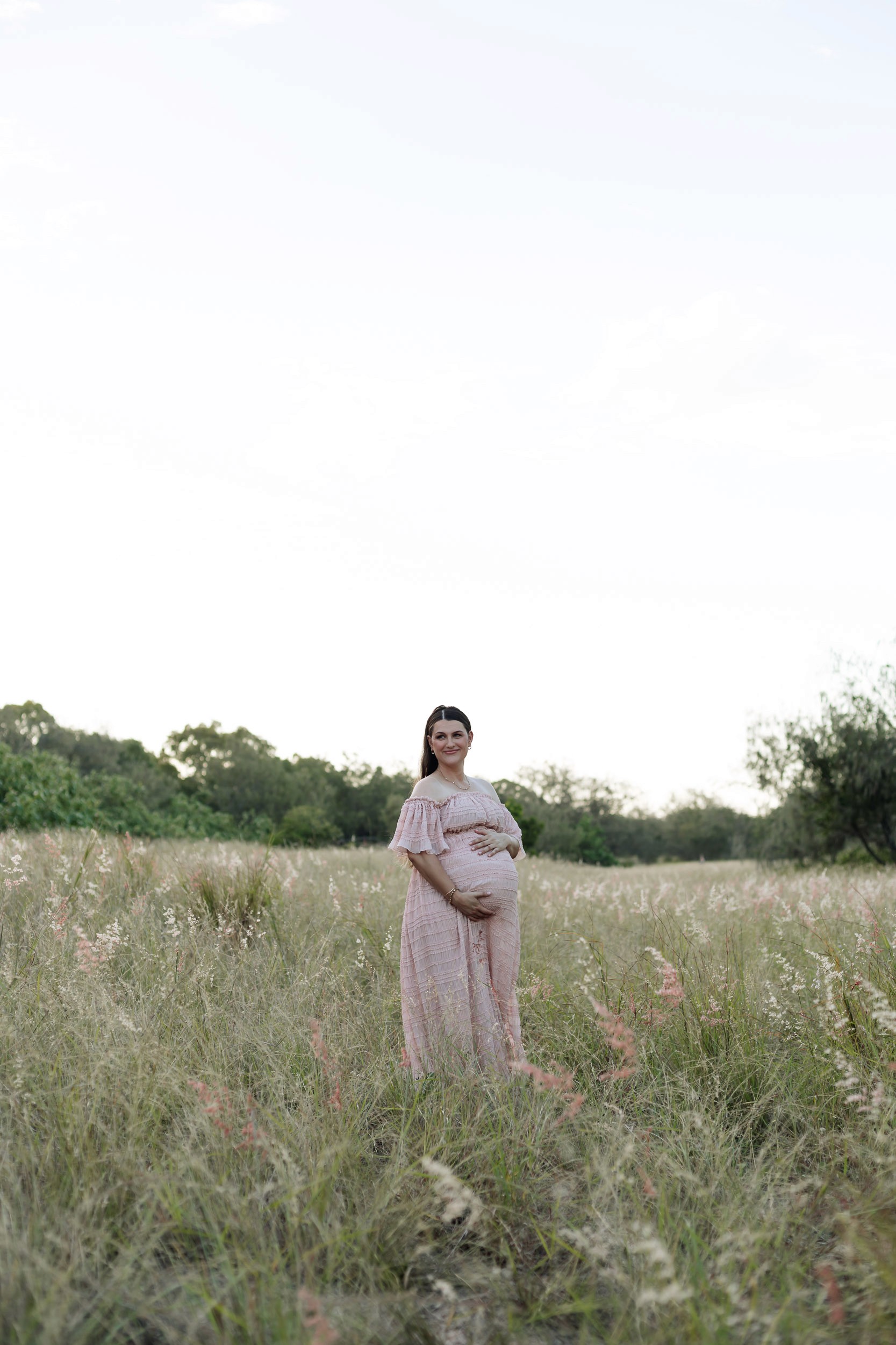 Pregnant woman standing in long golden grass at sunset wearing a flowing dress during Mackay maternity photography session