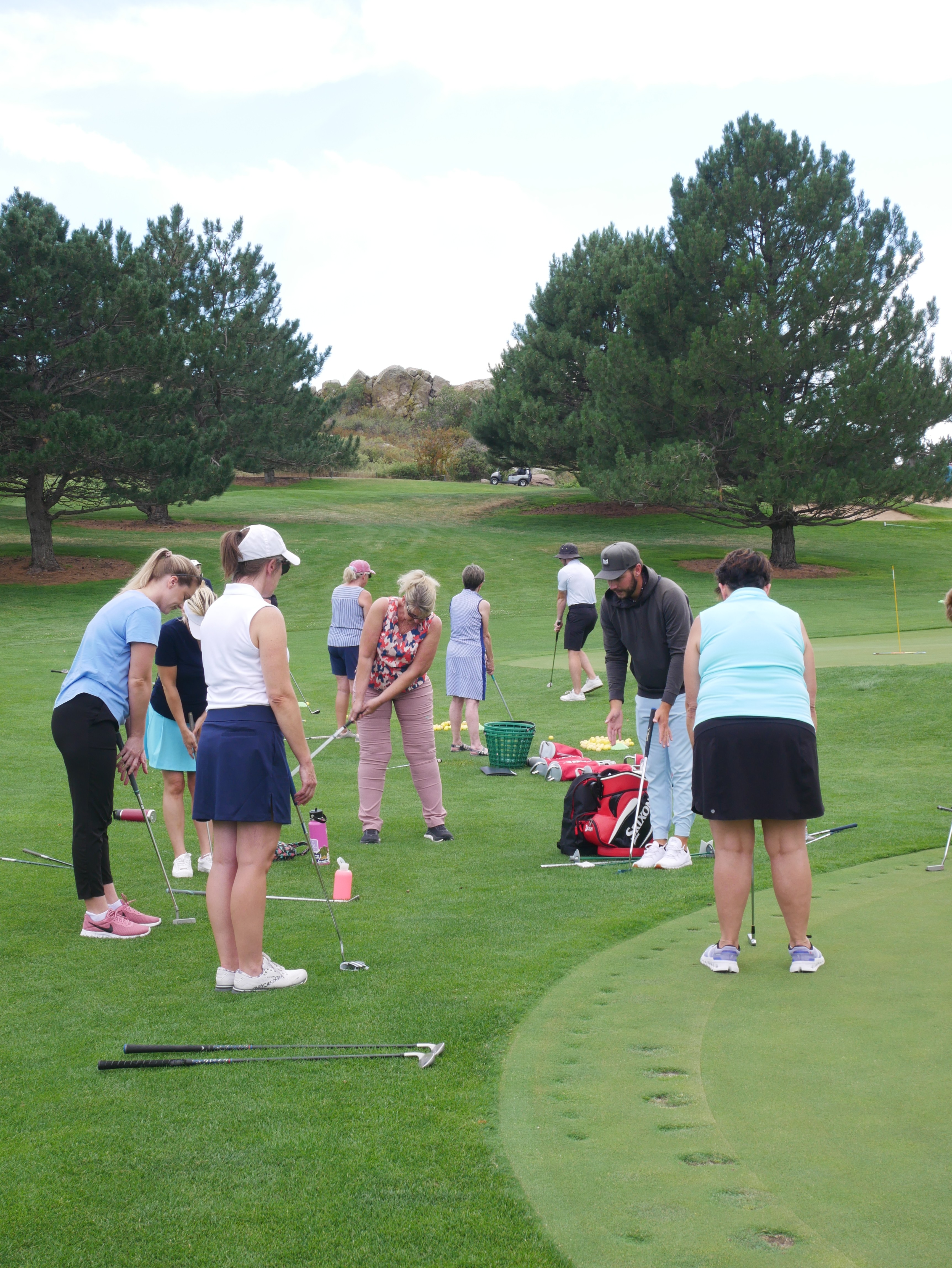 A group of students at the edge of a green, chipping balls towards net targets.