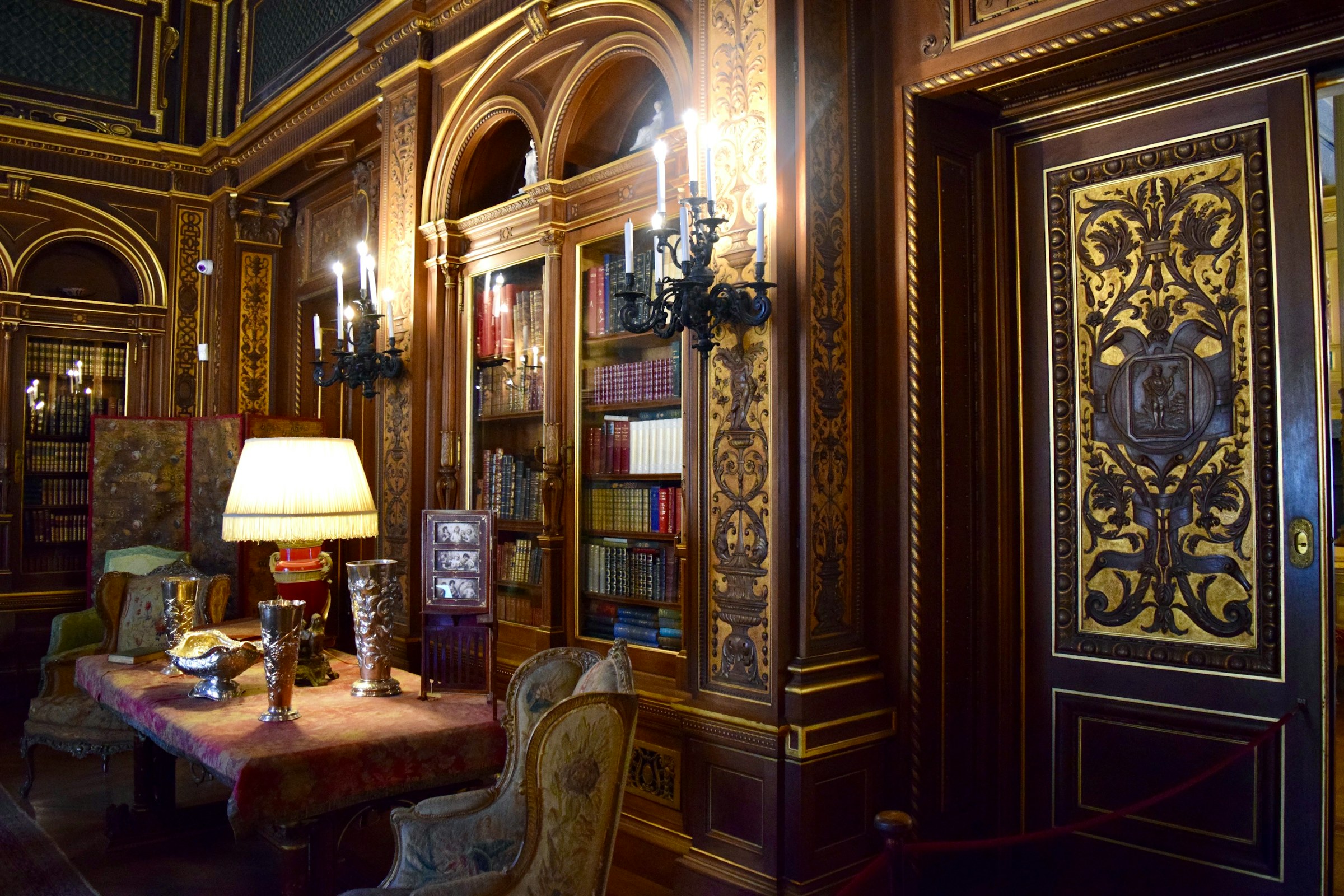 A dark, ornate study: carved wood panels, candle sconces, a bookcase and a shaded lamp on a table