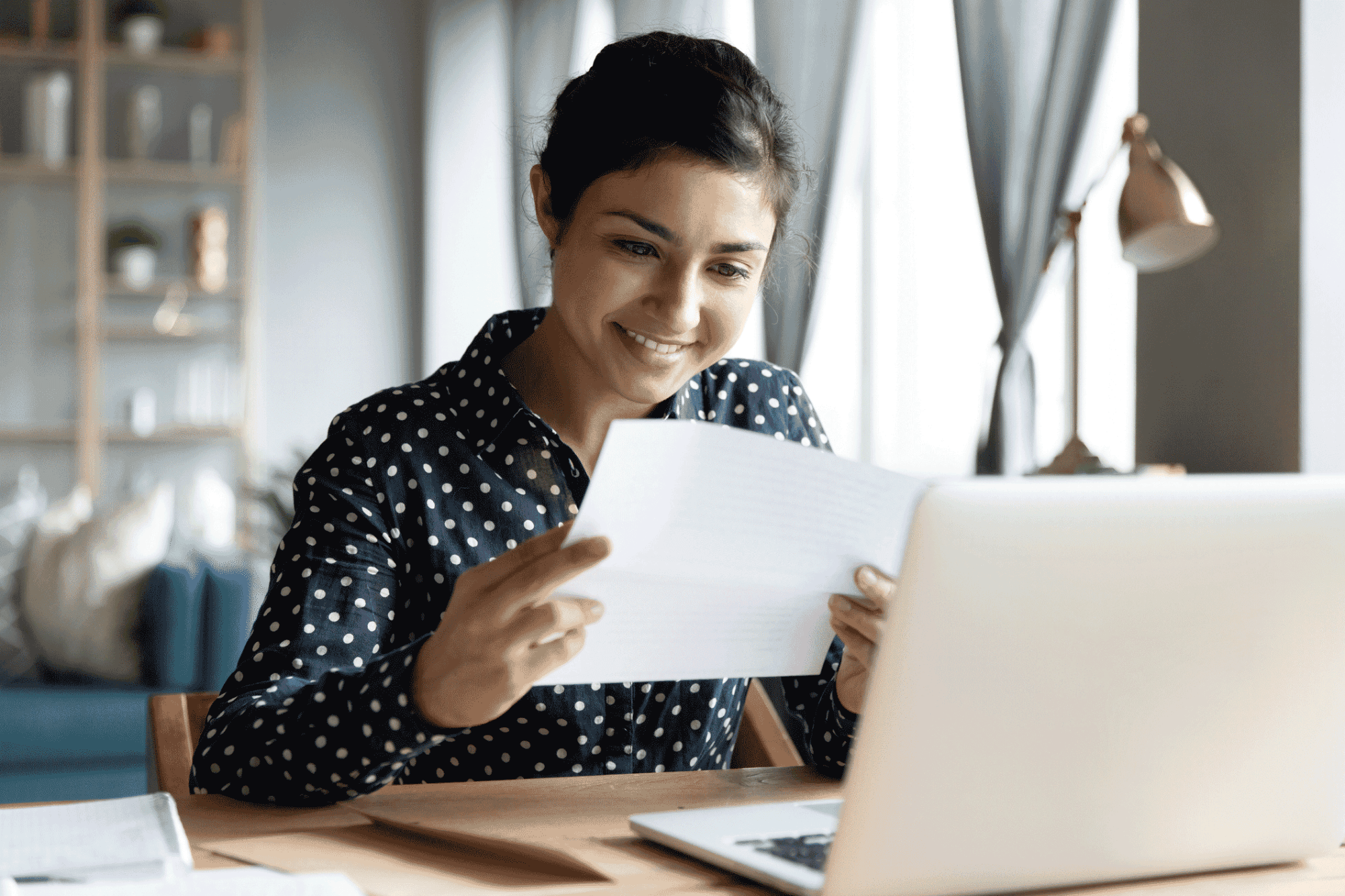 Woman at home smiling while reviewing paperwork next to a laptop.