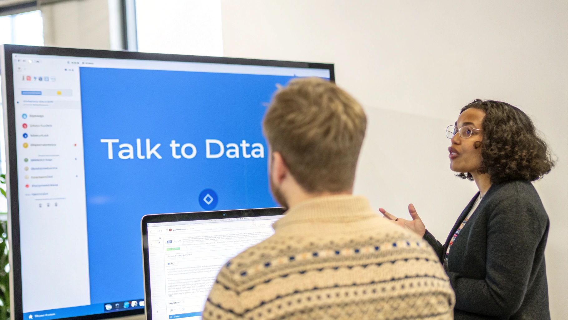 A diverse woman presents to a man in an office, with a screen showing 'Talk to Data'.