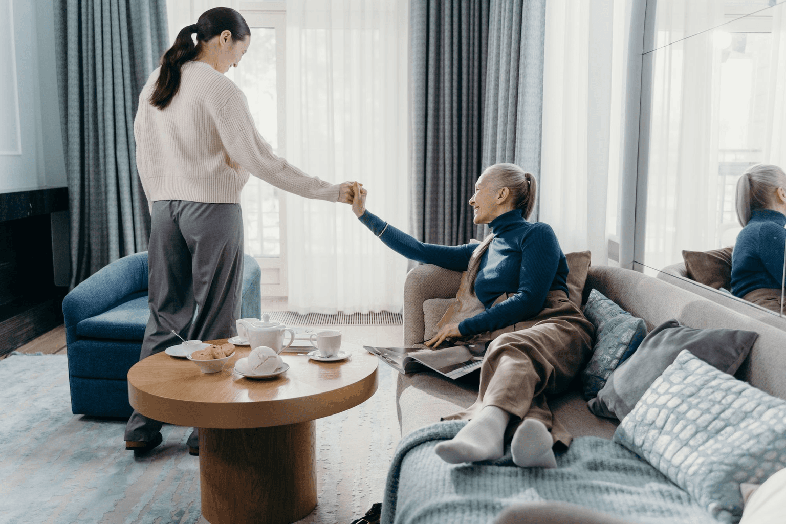 A daughter reaching for her mother's hand in her bright living room, tea on the table.