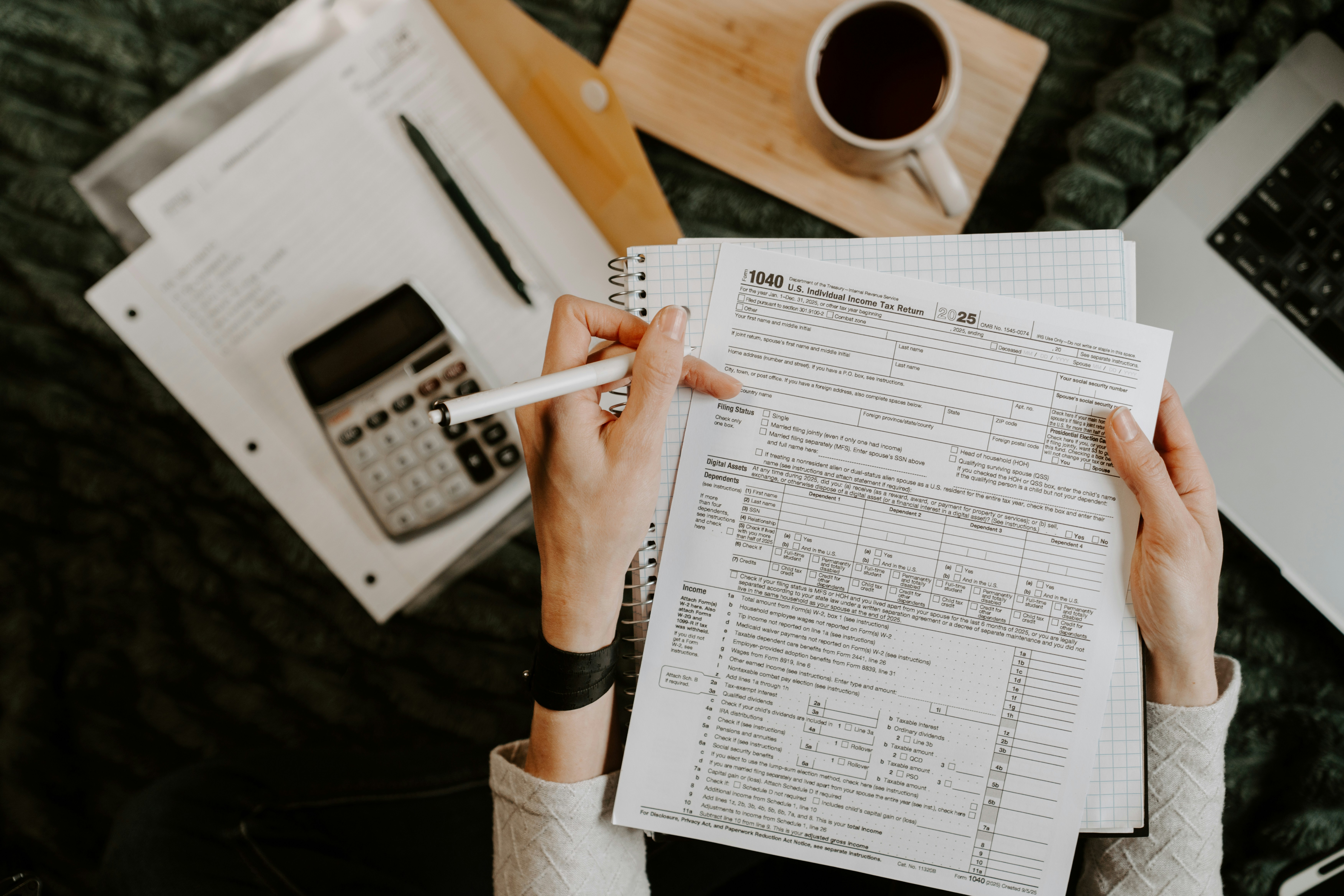 Person reviewing a tax form with a pen, calculator, notebook, and coffee on a desk.
