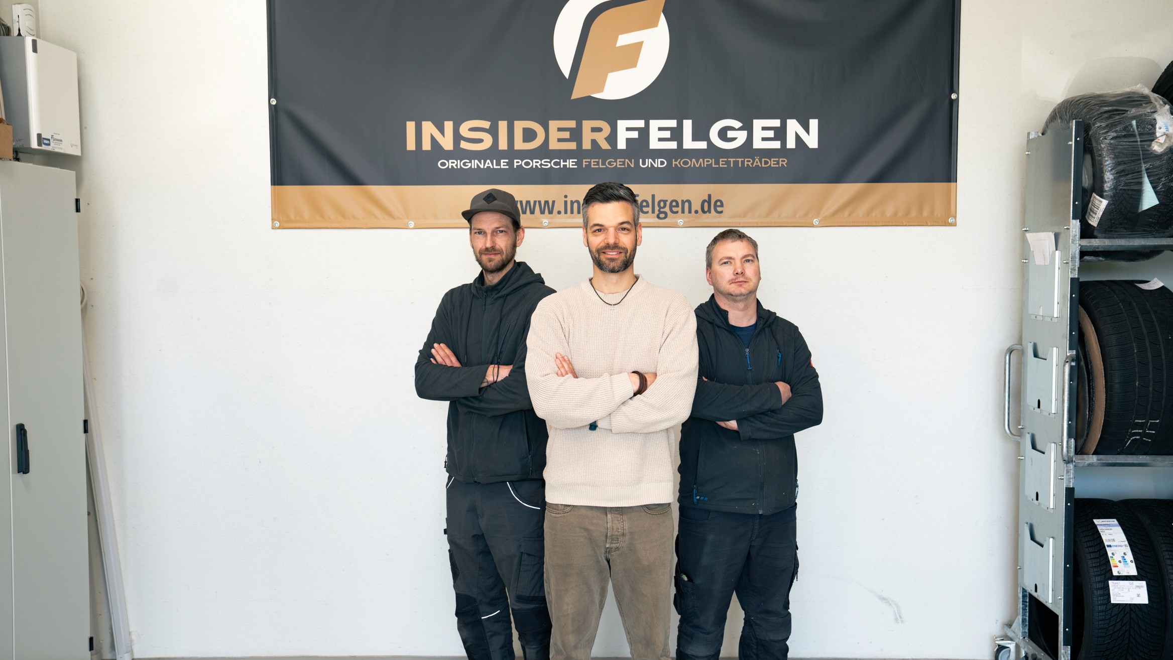 Schwarz-weiß Foto von drei Mitarbeitenden von Insiderfelgen, die mit verschränkten Armen vor einem Firmenbanner stehen – Teampräsentation eines spezialisierten Anbieters für originale Porsche Felgen.