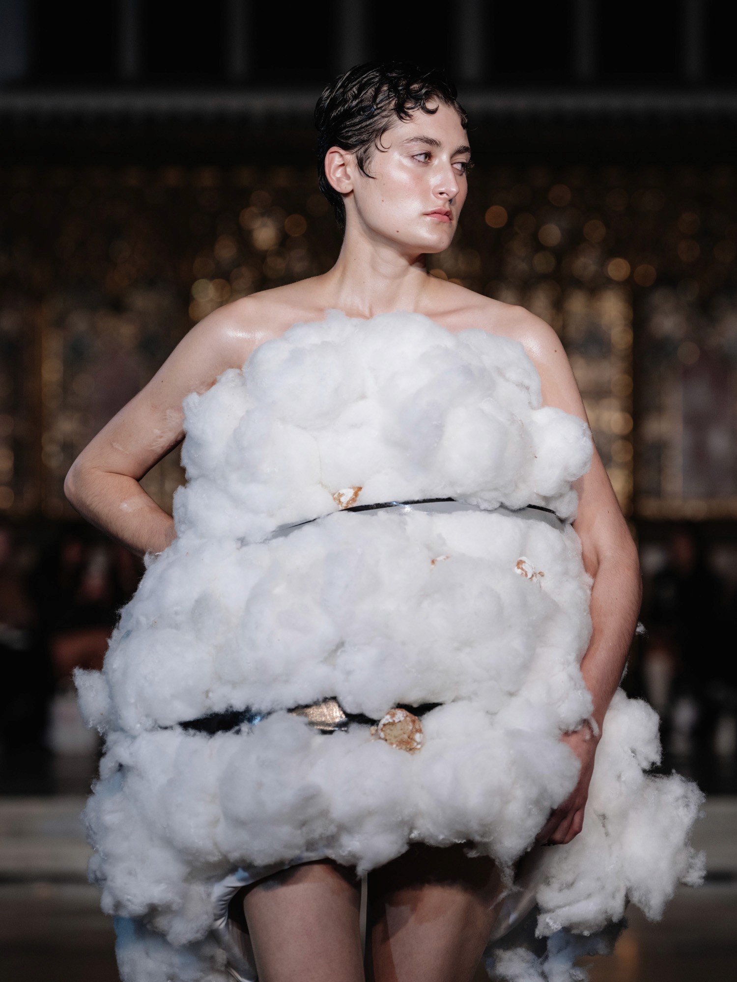 A model struts down the runway wearing an avant-garde dress resembling fluffy white clouds, set in an opulent venue with blurred figures in the background, highlighting fashion innovation and creativity.