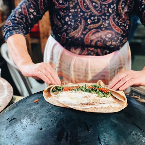 A person wearing a patterned shirt and apron folds a flatbread filled with greens over a round cooking surface.