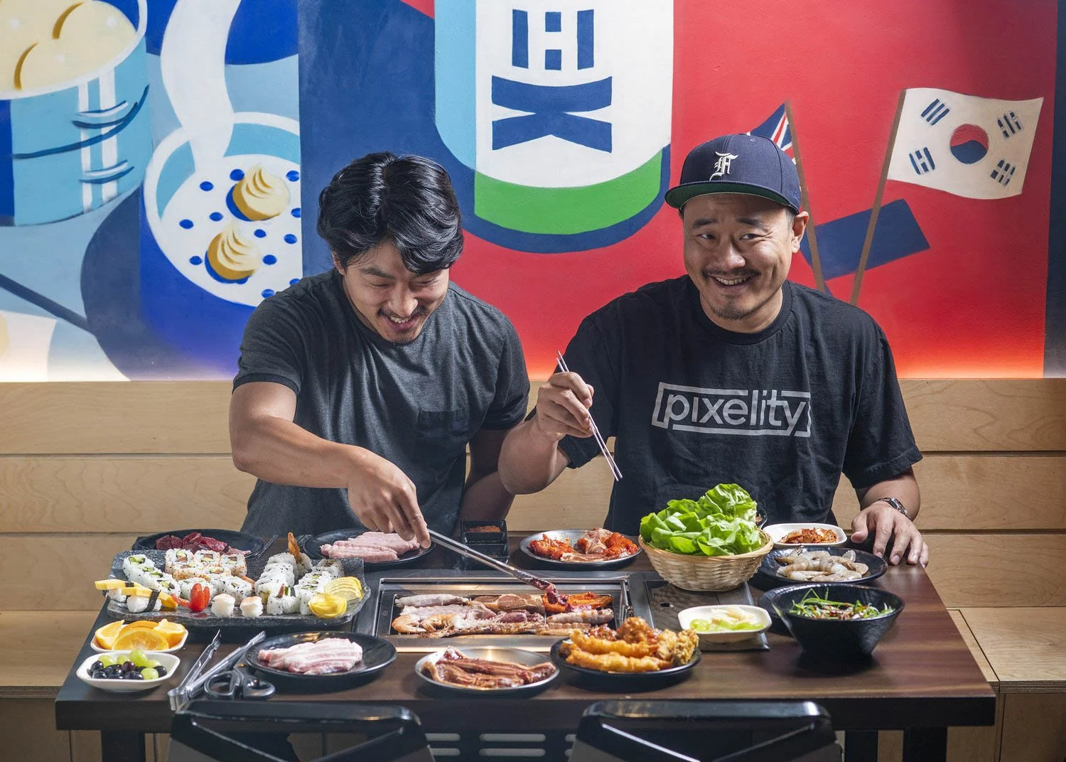 Two men prepare Korean BBQ at a table with grilled meats, sushi, and sides against a colorful backdrop featuring Korean flag and "pixelity" branding.