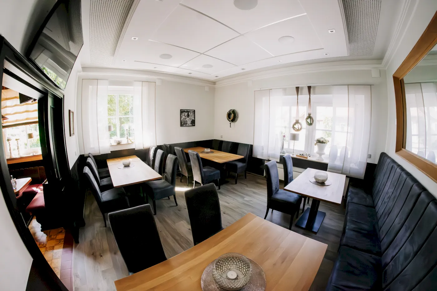 Perspektivische Aufnahme eines Gastraums mit Holztischen, schwarzen Stühlen und Sitzbänken, großem Spiegel und Fenster.