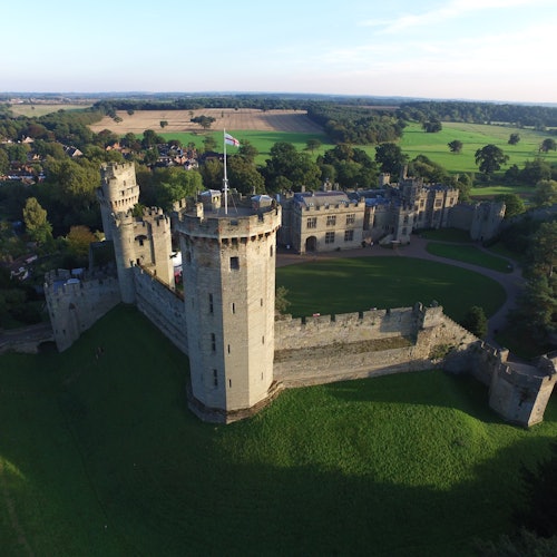 Ilmakuva historiallisesta linnasta, jossa on useita kivitorneja, vihreitä sisäpihoja sekä ympäröiviä peltoja ja puita.