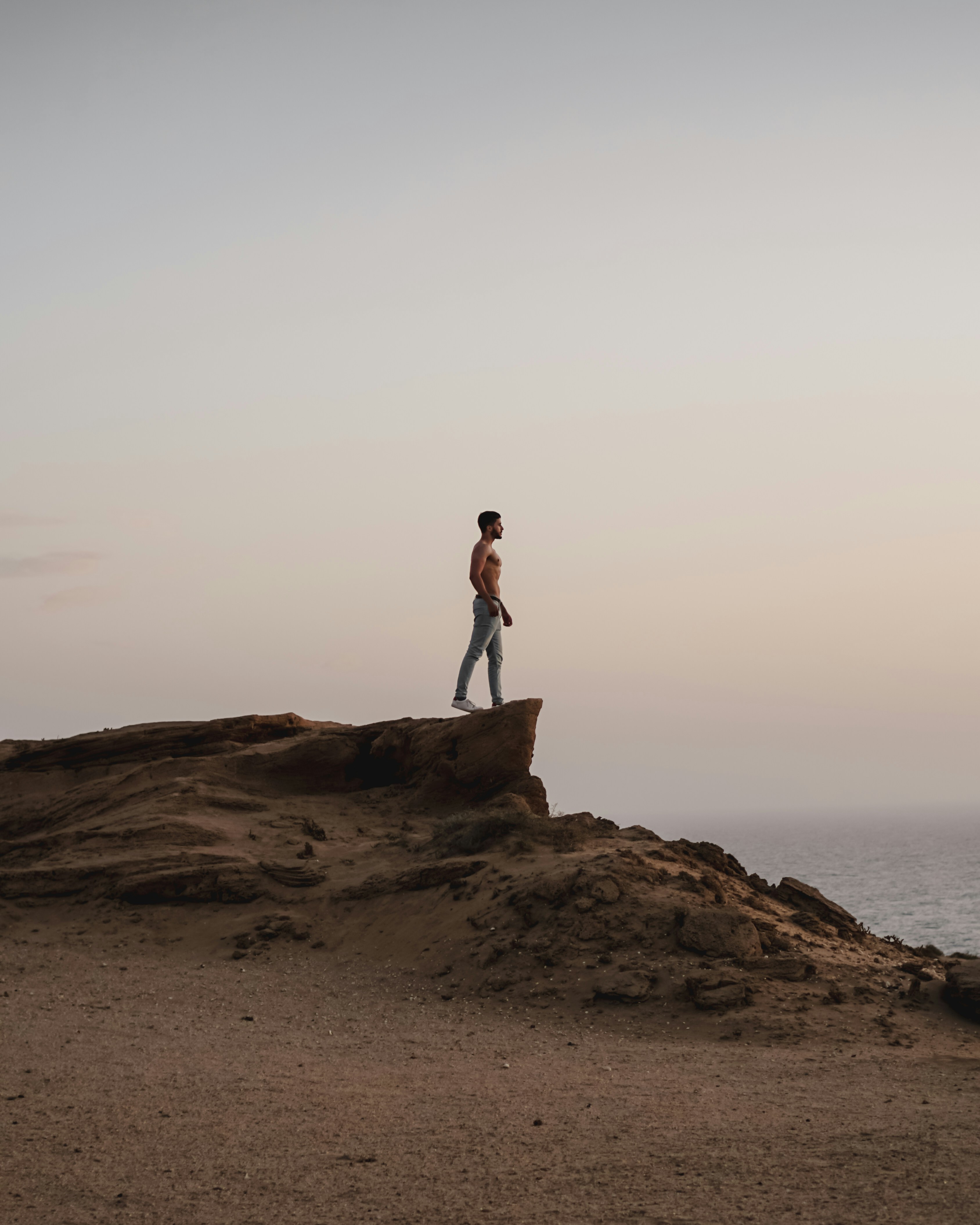 Person standing on a rocky coastal cliff overlooking the ocean at sunset.