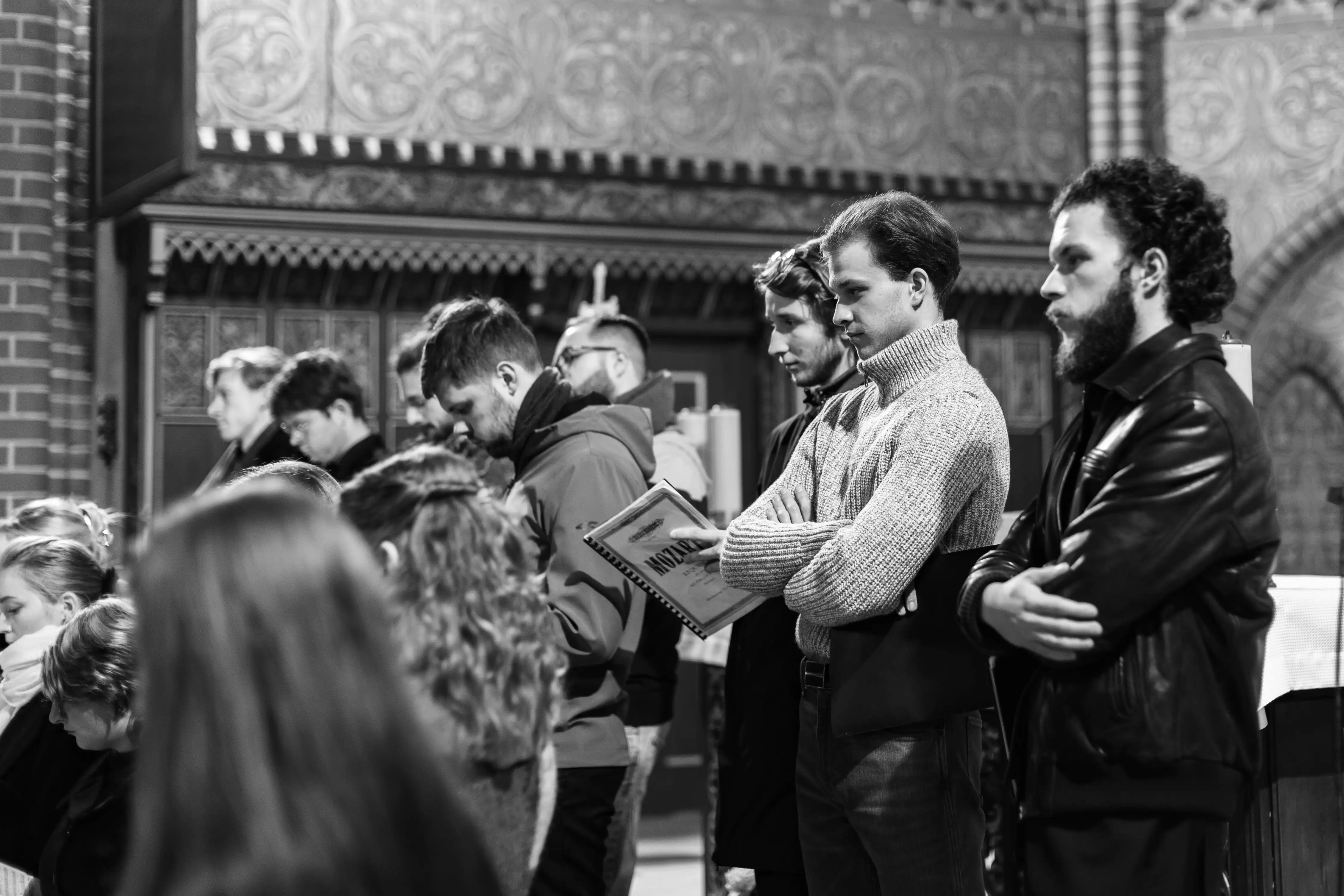 Grupa osób stoi w kościele podczas nabożeństwa, niektórzy trzymają modlitewniki. W tle widać ozdobne detale architektoniczne. Osoby są skupione i z zaangażowaniem uczestniczą w ceremonii.