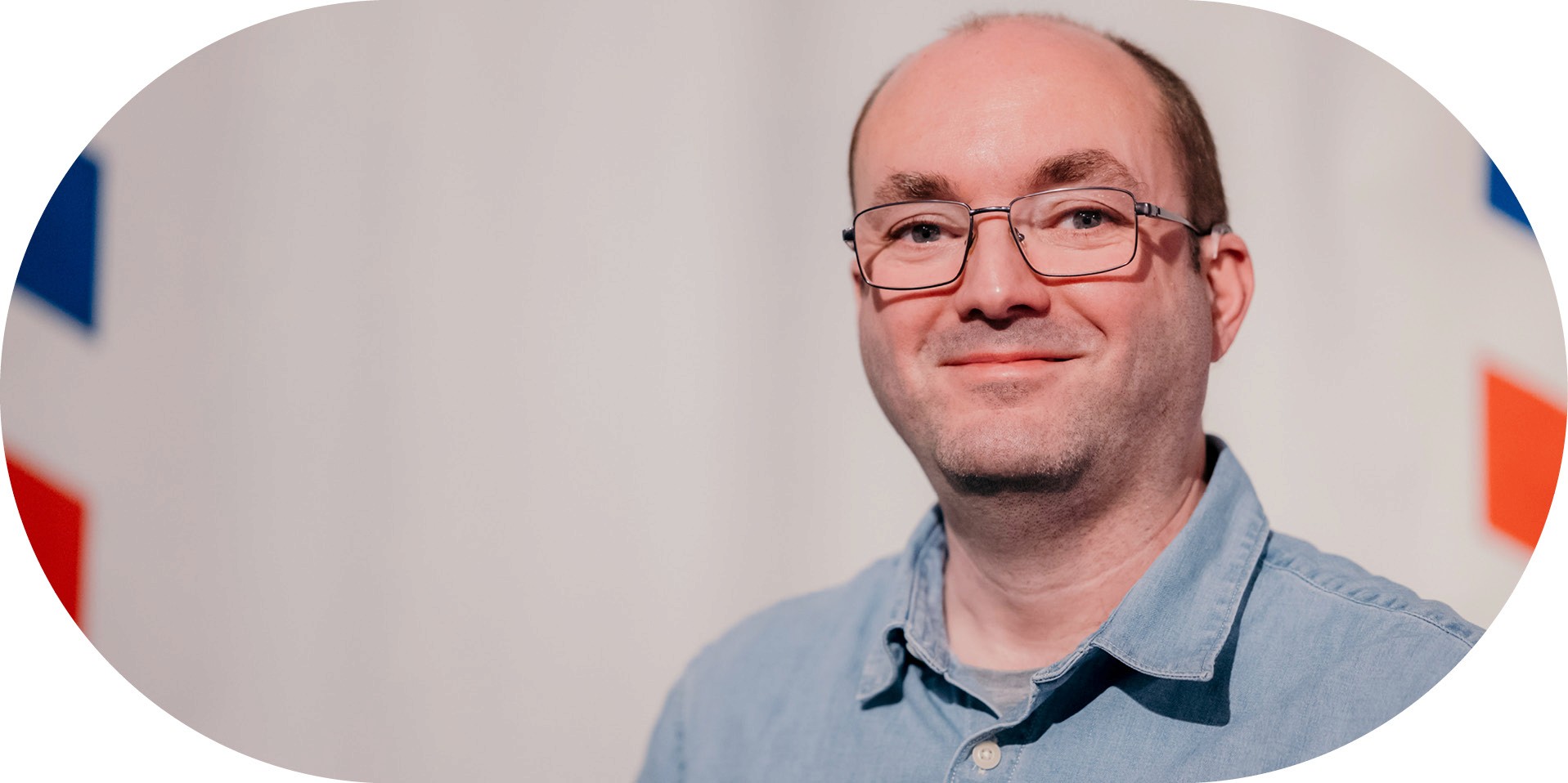 A man with short hair and glasses wearing a light blue shirt smiles in front of a white background
