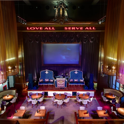 A stylish lounge with round tables and velvet chairs, a large screen, and illuminated signs reading "LOVE ALL" and "SERVE ALL."