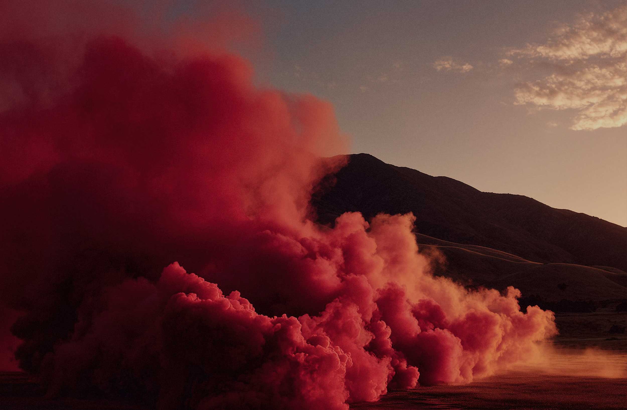 Bright red smoke plumes across a landscape of rolling hills at sunset.