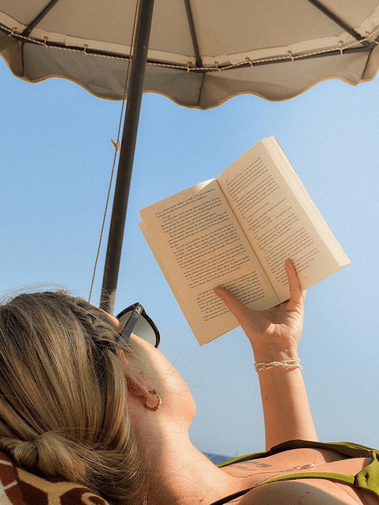 a woman laying under a beach umbrella wearing sunglasses while holding up the book she's reading with one hand