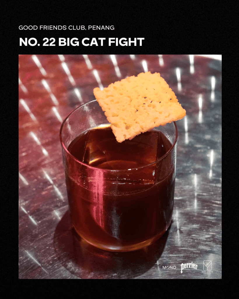 A close-up of a cocktail named 'Big Cat Fight' from Good Friends Club in Penang, featuring a glass with a dark drink and a savory cracker resting on top.
