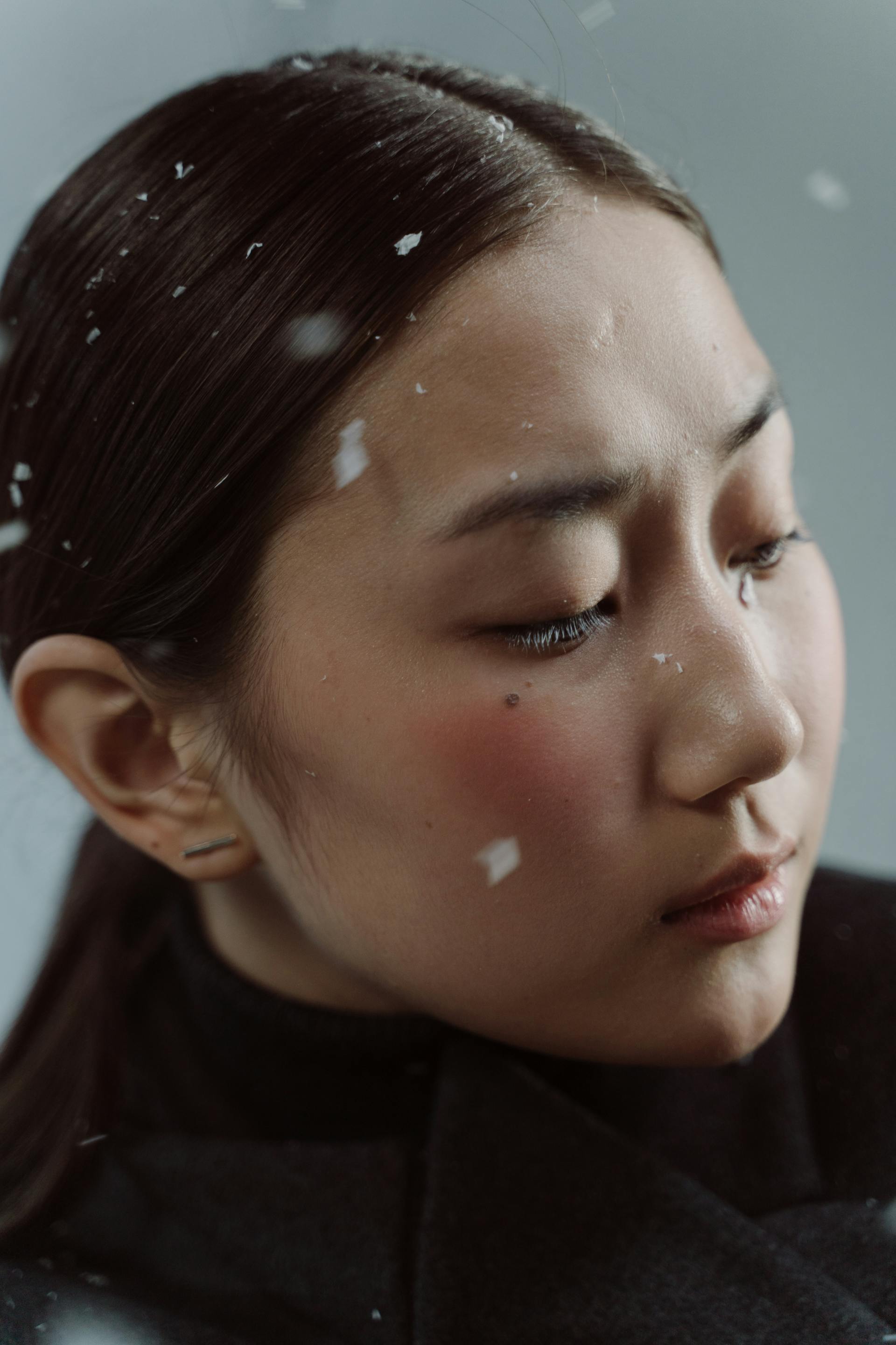 A close-up portrait of a woman looking downward with a somber expression as snowflakes rest on her dark hair and face.
