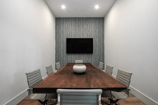 Modern conference room with a wooden table, chairs, and a screen on the wall. Bright, minimalist design.
