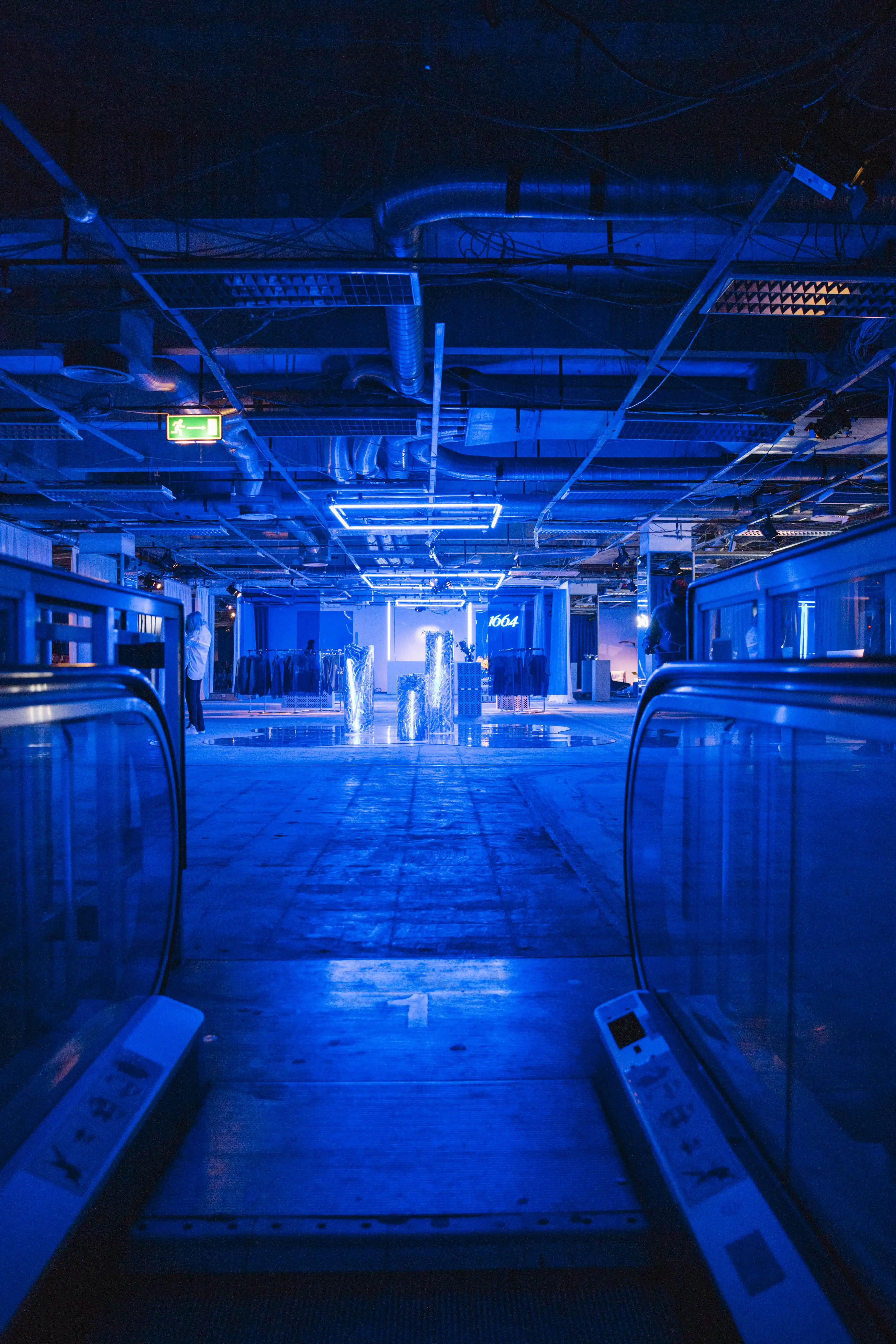 View down a frozen escalator into the blue-lit event level with neon frames and 1664 logo.