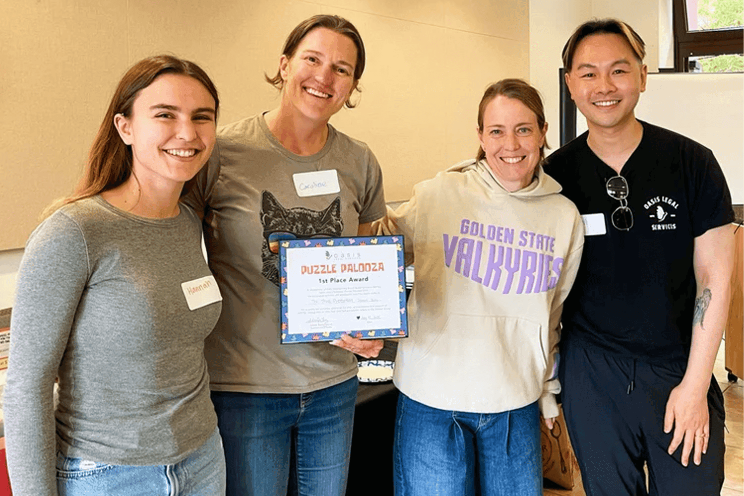 Quatro pessoas estão juntas em um ambiente interno, sorrindo e posando para uma foto em grupo, com uma pessoa segurando um certificado emoldurado por vencer o "Prêmio de 1º Lugar do Puzzle Palooza", demonstrando um senso de conquista e trabalho em equipe.
