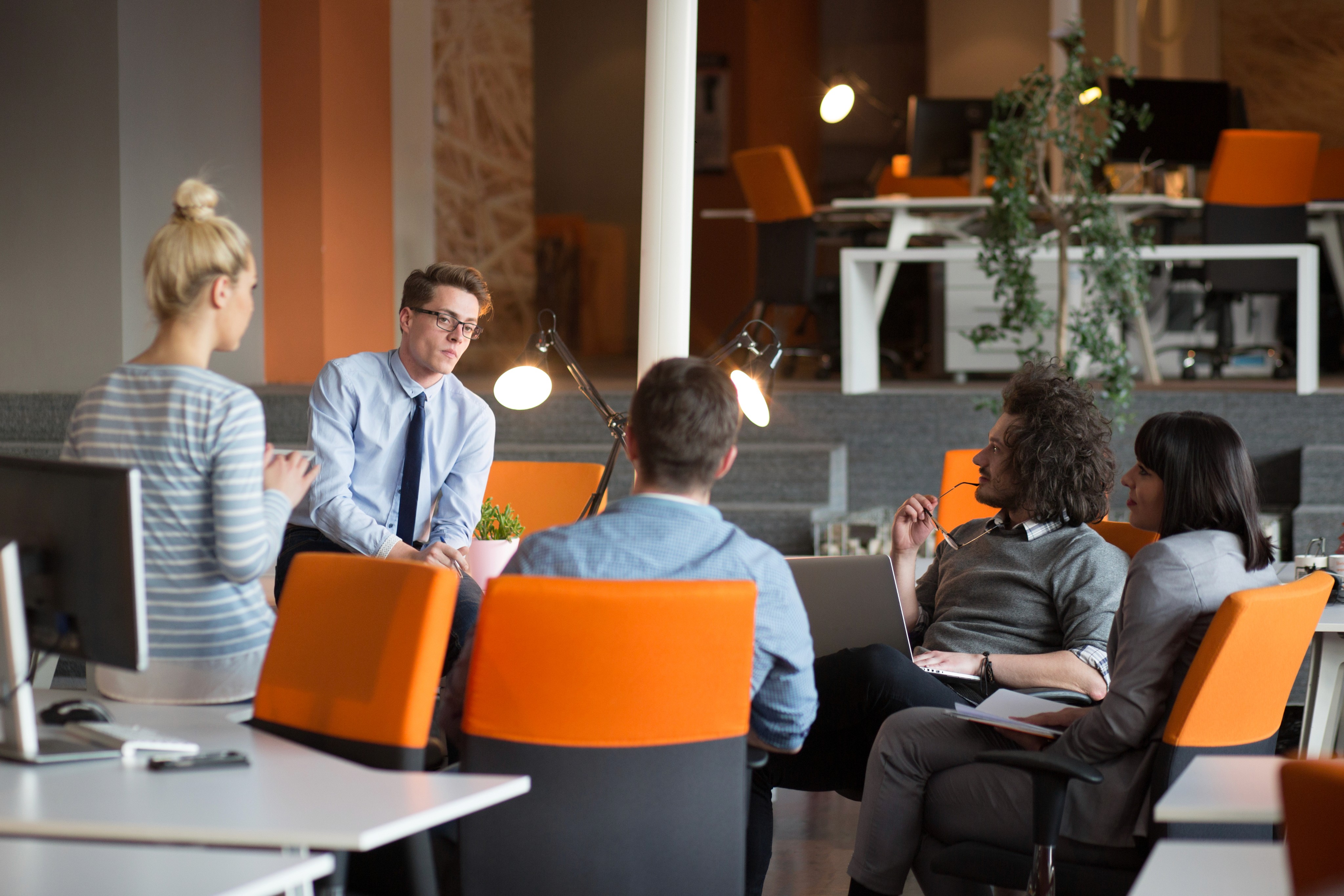Team of five coworkers having a discussion in a modern office with orange chairs.