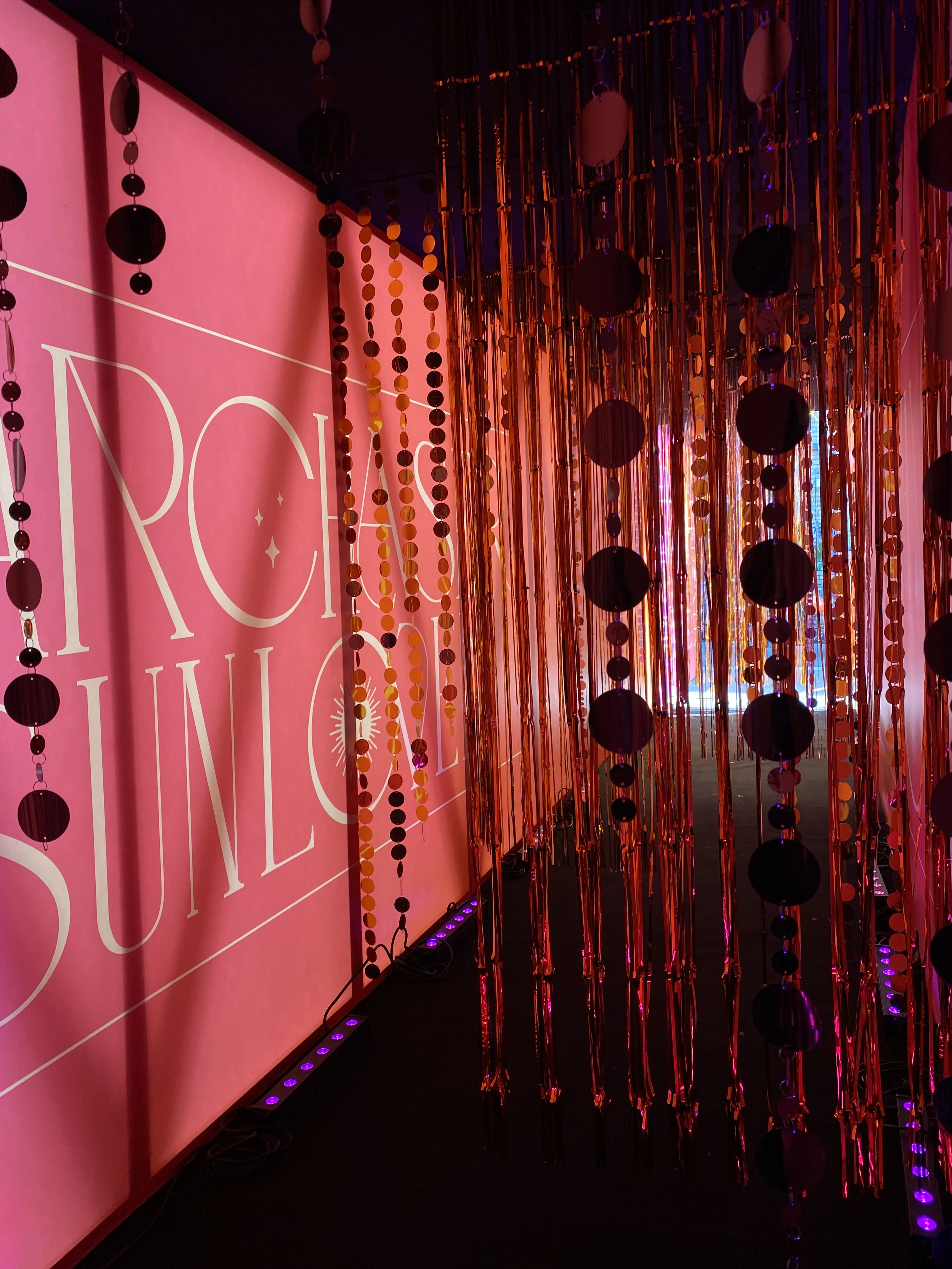 Pink-lit corridor with glossy fringe curtains, sequin garlands and illuminated lettering wall.