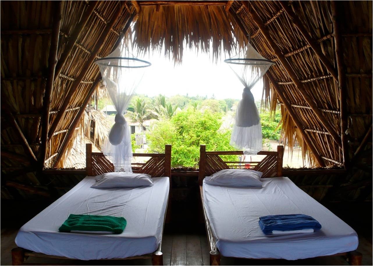 Dos camas con sábanas blancas y mosquiteros dentro de un loft rústico y al aire libre bajo un techo de paja.