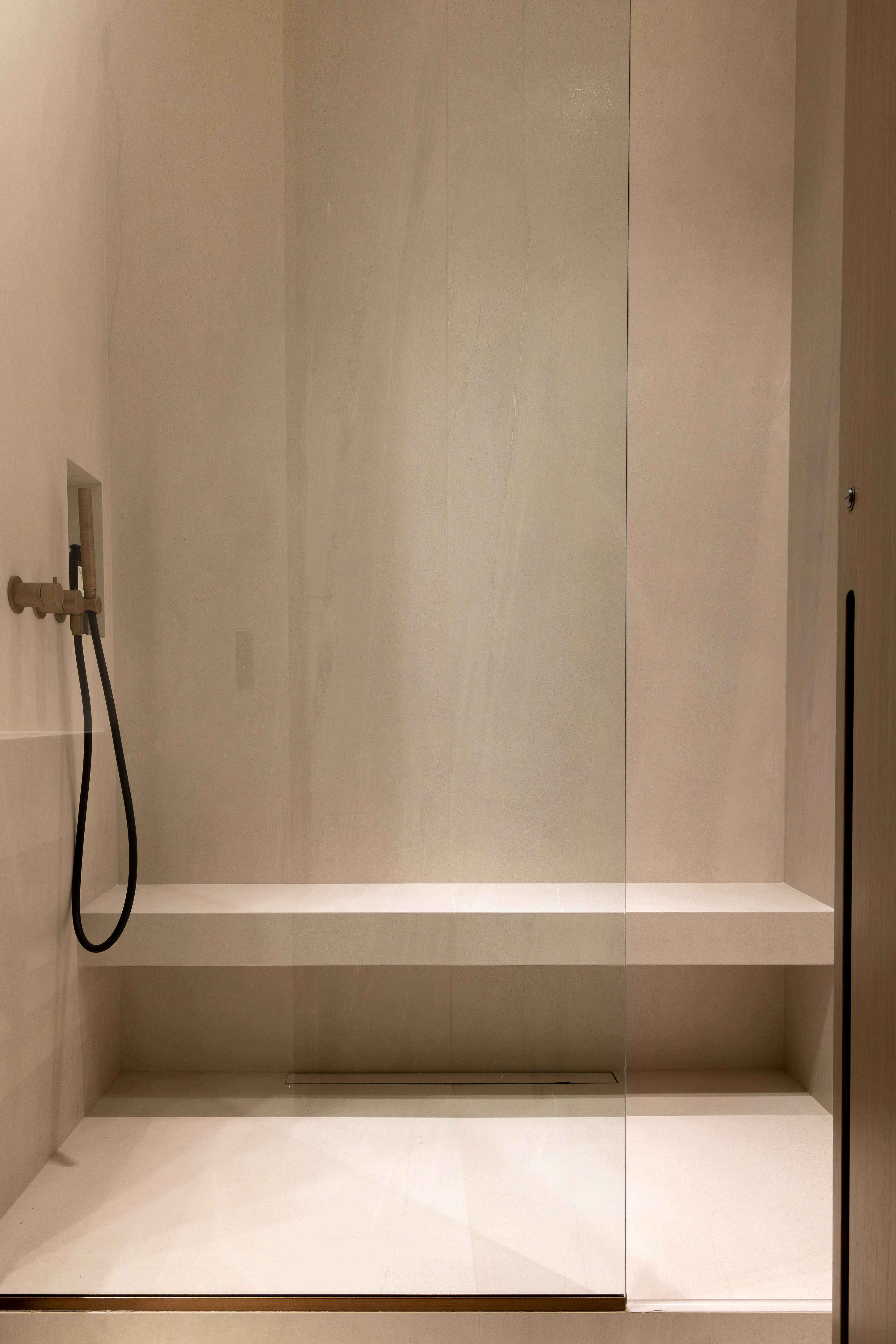 Douche italienne minimaliste en pierre claire avec douchette doree, banc integre et paroi vitree