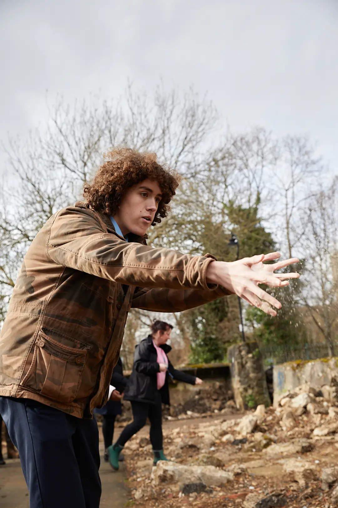 A person wearing a brown coat is aiming a firearm, while another individual is visible in the background.
