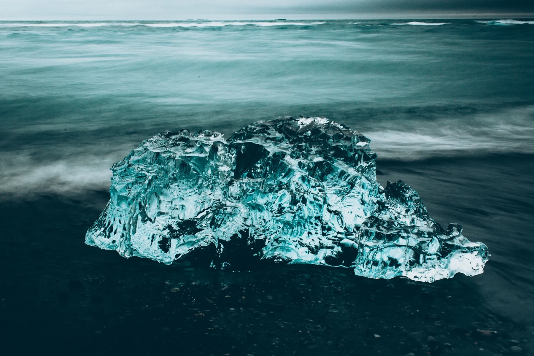 A large clear iceberg stranded on black volcanic sand as surf washes past