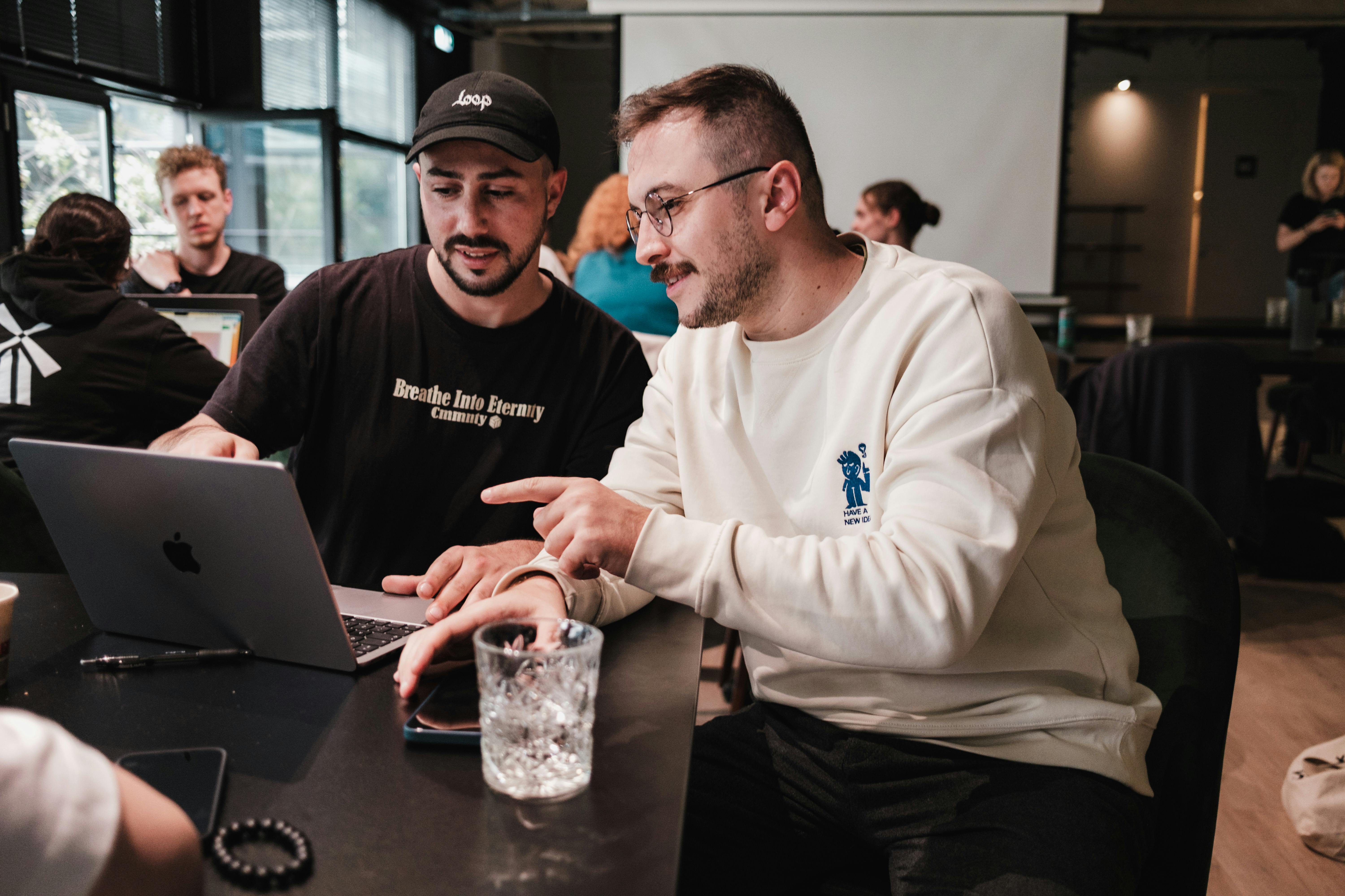 Two men working together on a laptop during a casual meeting, discussing ideas with other people in the background.